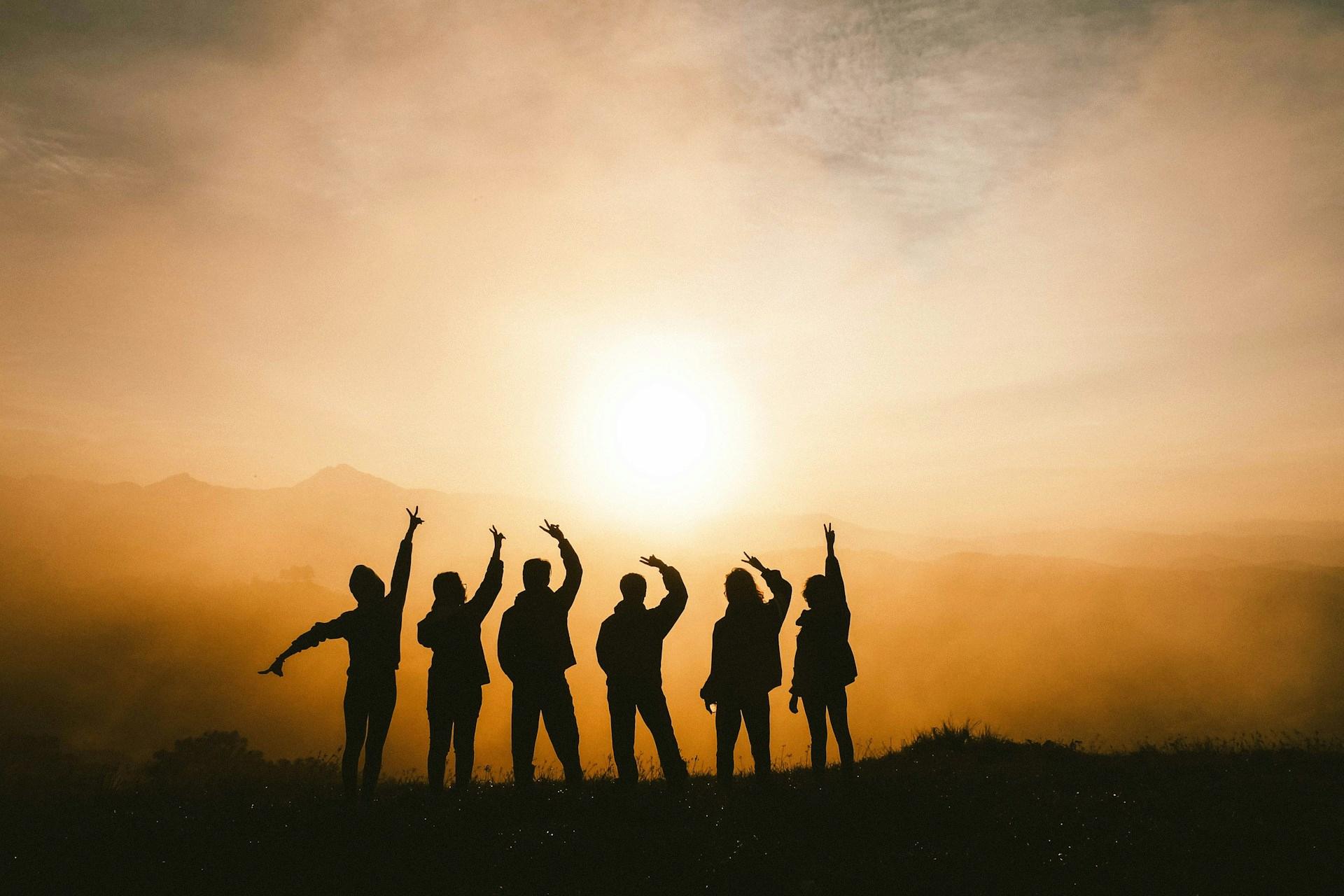 A group of people are silhouetted against a sunset, with their arms raised in a gesture of celebration or joy. The warm colors of the sky and the silhouettes create a peaceful and celebratory atmosphere.