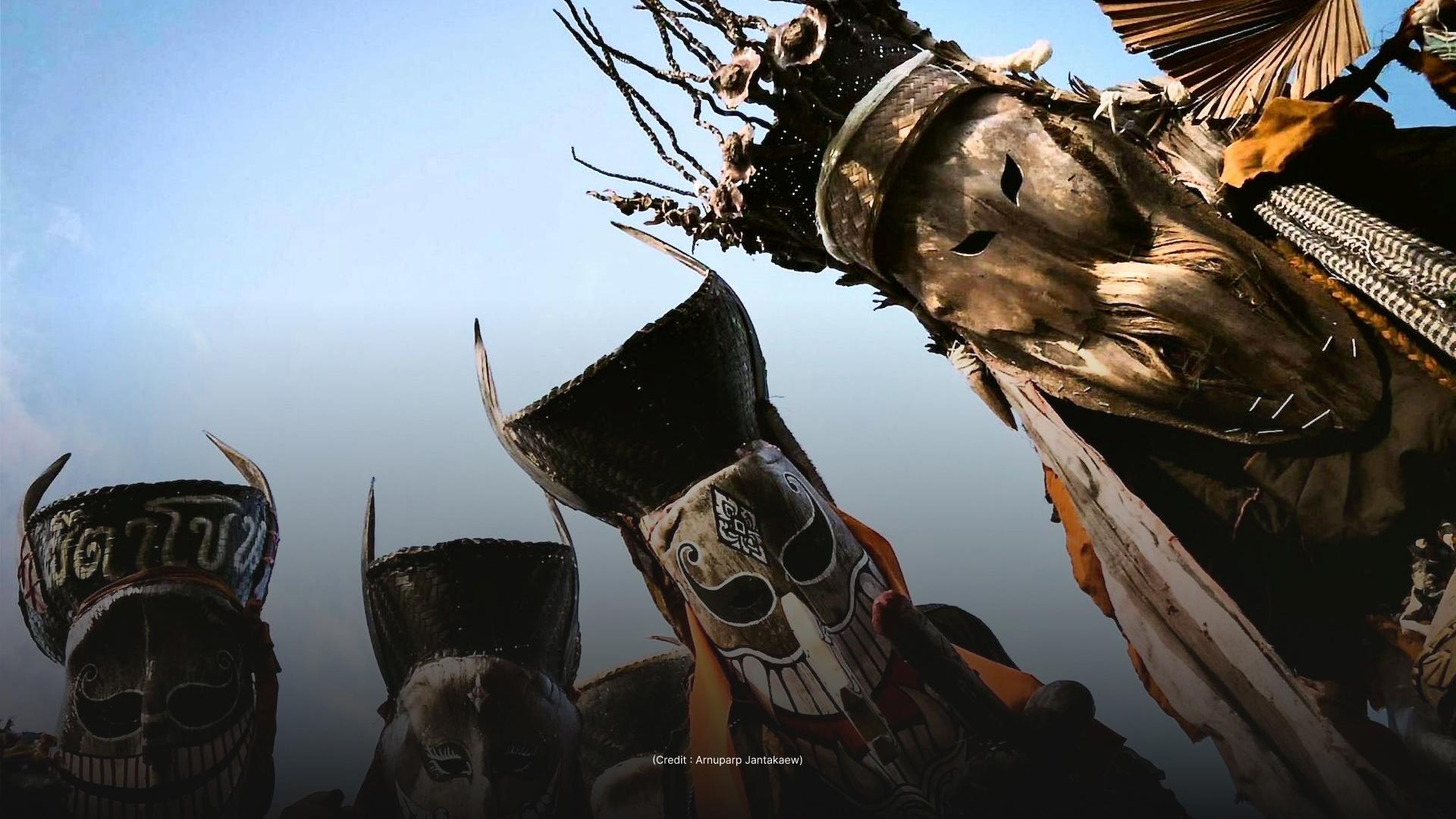 The image shows a low-angle view of several elaborate masks, likely used in a cultural ceremony. The masks are made from wood and other natural materials, and are set against a bright sky.