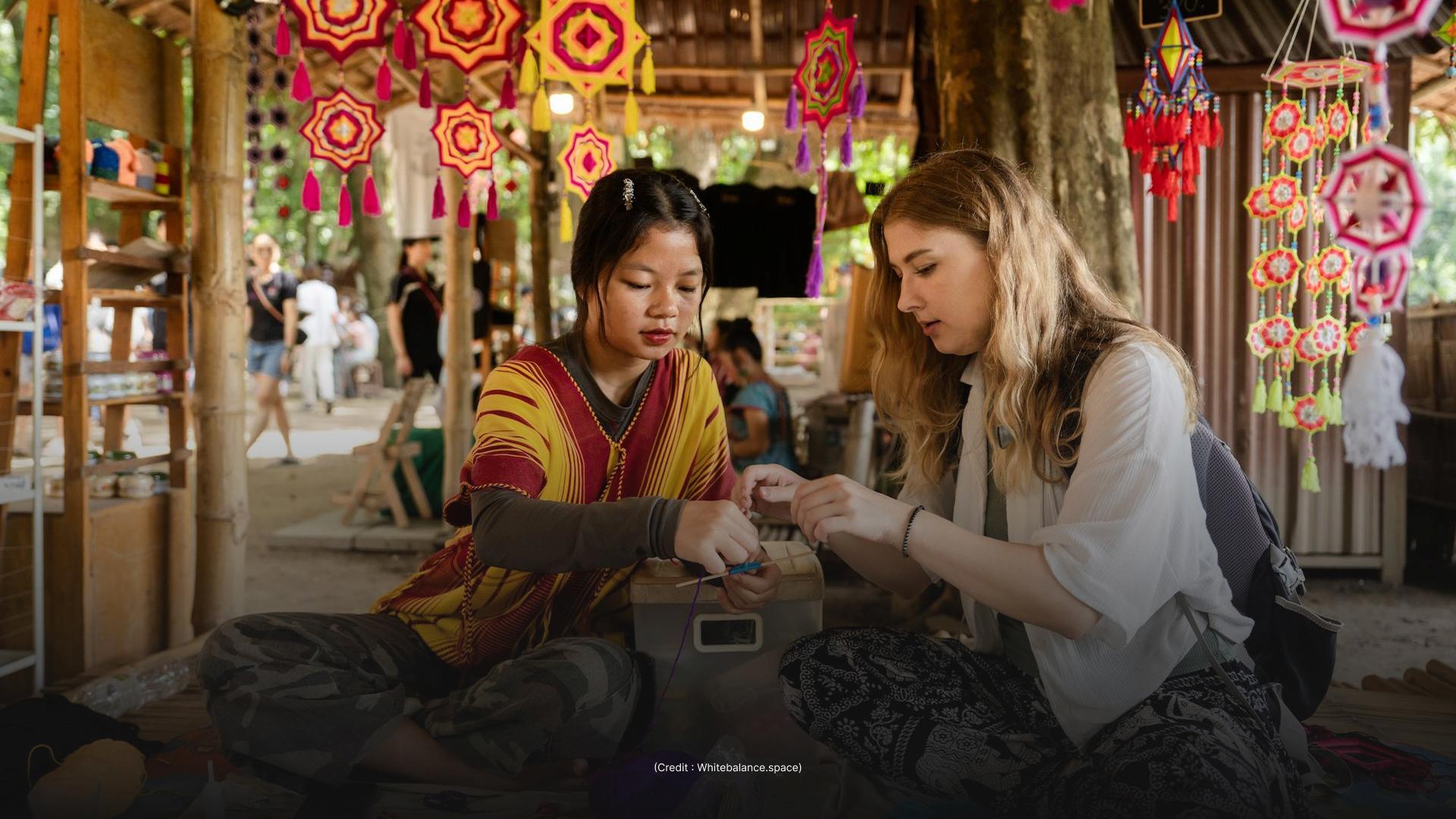 Two women are seated on the ground, crafting with small tools. They are surrounded by colorful hanging decorations and appear to be in a market or workshop setting.