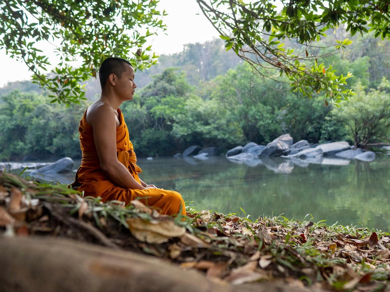 A Buddhist monk in orange robes meditates by a tranquil river. The serene scene is framed by lush greenery and a backdrop of trees.