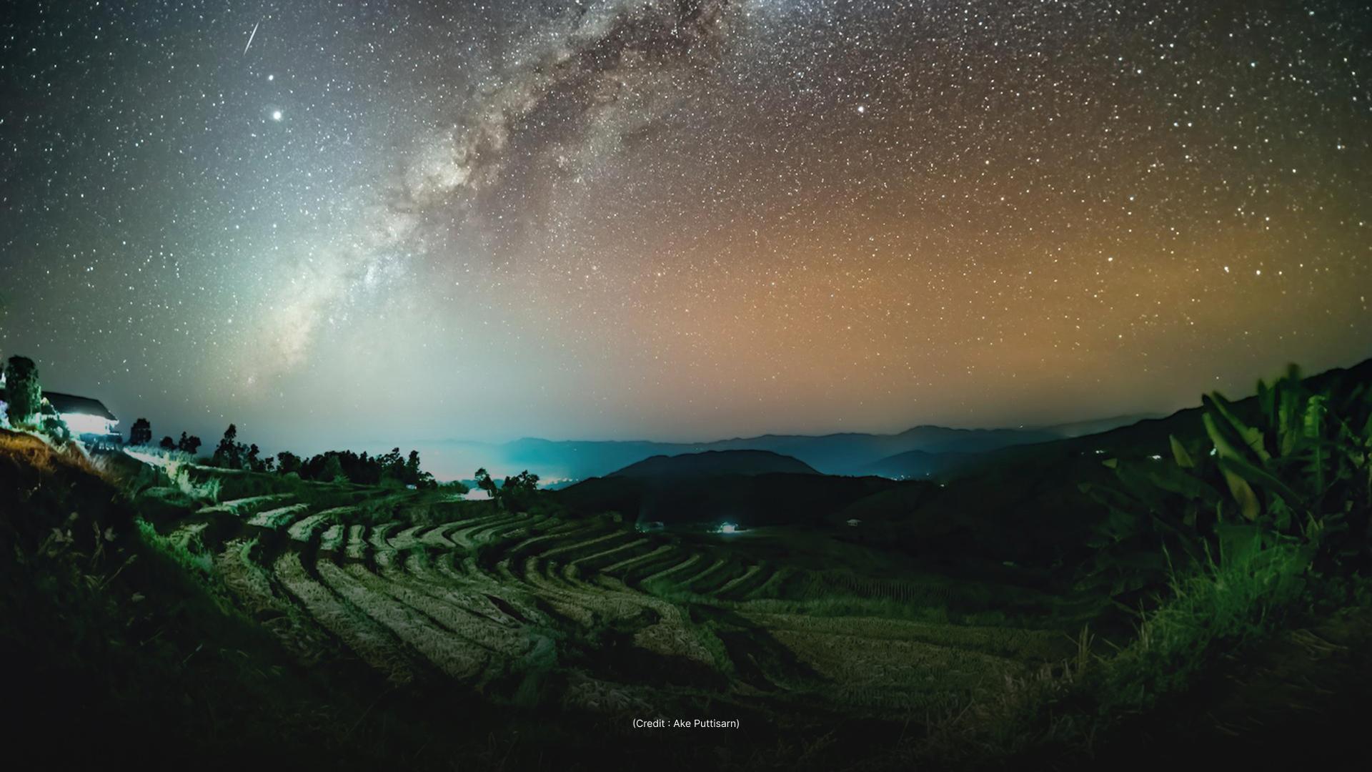 The image depicts a night scene of a terraced landscape under a starry sky, with the Milky Way prominently visible. The foreground features the terraced fields and some vegetation, while the background shows a range of dark mountains.